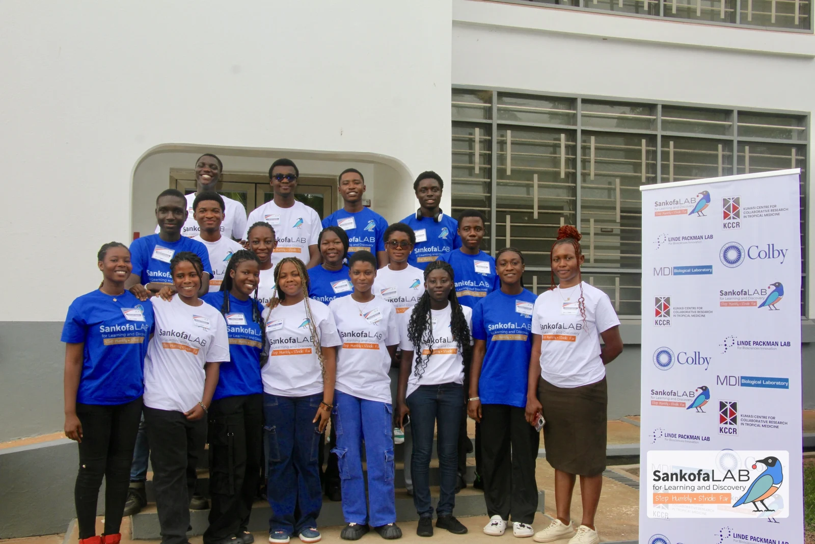 The 2026 Sankofa STEM Fellows stand in three rows in Sankofa Lab shirts outside the research centre, beside a banner listing the fellowship's partners