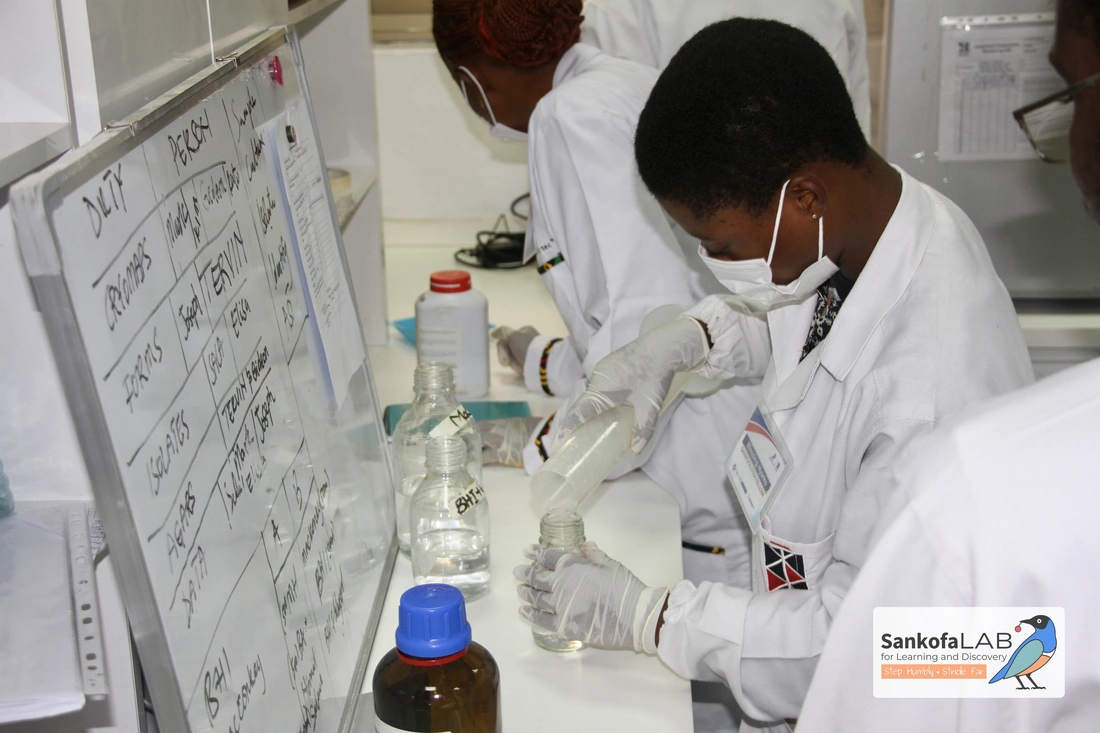 A fellow in a lab coat and mask pours liquid into a glass bottle beside a whiteboard listing the day's duties and the fellows assigned to them