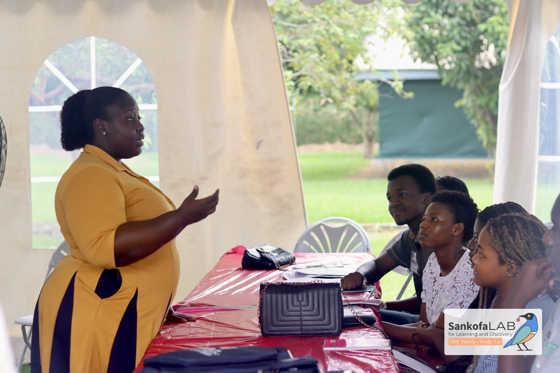 A speaker stands and addresses fellows seated along a long table under a tent